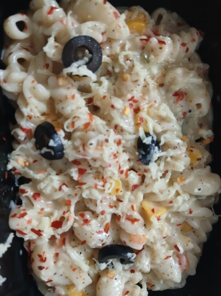 A close-up of delicious, creamy white sauce pasta loaded with melted cheese, red chili flakes, corn, and black olives.